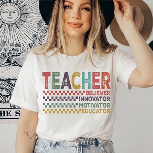 Peut inclure: Un t-shirt blanc avec le mot "TEACHER" en lettres r&eacute;tro color&eacute;es. Sous le mot, il y a quatre lignes de texte qui disent "BELIEVER", "INNOVATOR", "MOTIVATOR" et "EDUCATOR".