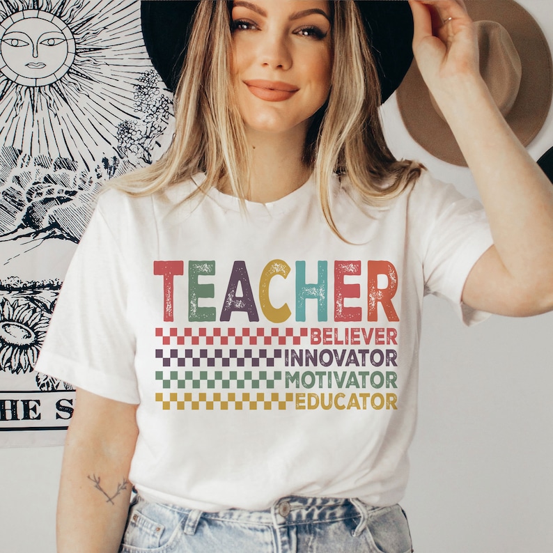 Peut inclure: Un t-shirt blanc avec le mot "TEACHER" en lettres r&eacute;tro color&eacute;es. Sous le mot, il y a quatre lignes de texte qui disent "BELIEVER", "INNOVATOR", "MOTIVATOR" et "EDUCATOR".