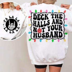 Peut inclure: Sweat-shirt blanc avec un motif de Noël au dos qui dit "Deck the halls and not your husband" avec une boule rouge et des guirlandes lumineuses. Le devant du sweat-shirt a un motif noir d'une main de squelette avec le texte "Fa La La La La La" autour.