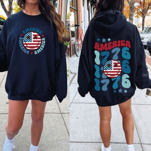 Pode incluir: Duas sweatshirts pretas com tema patri&oacute;tico. Uma sweatshirt apresenta um rosto sorridente com a bandeira americana e a palavra "AMERICA" repetida ao redor do rosto. A outra sweatshirt apresenta a palavra "AMERICA" com o ano "1776" repetido em um padr&atilde;o azul e verde.