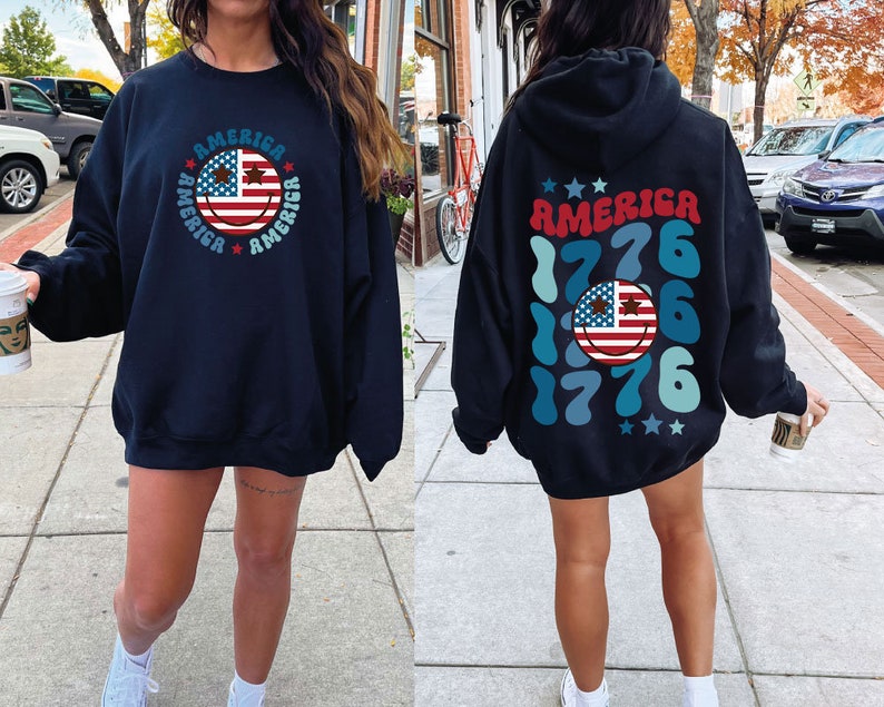Pode incluir: Duas sweatshirts pretas com tema patri&oacute;tico. Uma sweatshirt apresenta um rosto sorridente com a bandeira americana e a palavra "AMERICA" repetida ao redor do rosto. A outra sweatshirt apresenta a palavra "AMERICA" com o ano "1776" repetido em um padr&atilde;o azul e verde.