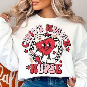 Op de afbeelding: Witte trui met een rode hartgrafiek waarop in roze en rode letters "Cupid's Favorite Nurse" staat. Het hart draagt een verpleegstersmuts en houdt een spuit en een stethoscoop vast.