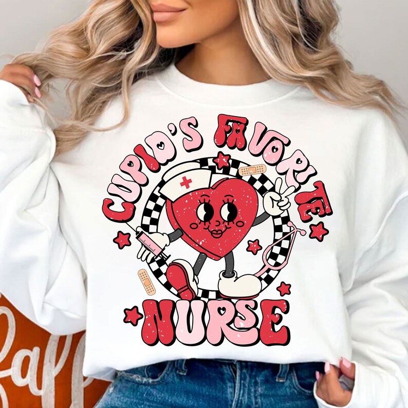 Op de afbeelding: Witte trui met een rode hartgrafiek waarop in roze en rode letters "Cupid's Favorite Nurse" staat. Het hart draagt een verpleegstersmuts en houdt een spuit en een stethoscoop vast.