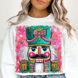 K&ouml;nnte beinhalten: Wei&szlig;es Sweatshirt mit einer farbenfrohen, k&uuml;nstlerischen Illustration eines Nussknackers. Der Nussknacker hat einen t&uuml;rkisfarbenen Hut mit einer rosa Schleife, einen schwarzen Schnurrbart und einen wei&szlig;en Bart. Der Hintergrund ist rosa mit wei&szlig;en und goldenen Sternen.
