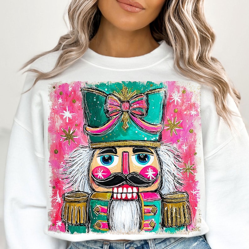 K&ouml;nnte beinhalten: Wei&szlig;es Sweatshirt mit einer farbenfrohen, k&uuml;nstlerischen Illustration eines Nussknackers. Der Nussknacker hat einen t&uuml;rkisfarbenen Hut mit einer rosa Schleife, einen schwarzen Schnurrbart und einen wei&szlig;en Bart. Der Hintergrund ist rosa mit wei&szlig;en und goldenen Sternen.