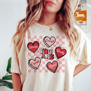May include: A white t-shirt with a pink and white checkered background and pink hearts with positive affirmations. The hearts say "Strong", "Enough", "Worthy", "Capable", and "Loved". The text "You Are" is in large red letters.