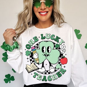 Pu&ograve; includere: Felpa bianca con un cartone animato di trifoglio verde che tiene una matita e una mela. Il testo "One Lucky Teacher" &egrave; scritto in un cerchio verde attorno al trifoglio.