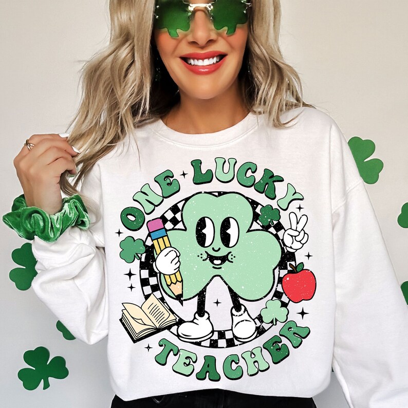 Pu&ograve; includere: Felpa bianca con un cartone animato di trifoglio verde che tiene una matita e una mela. Il testo "One Lucky Teacher" &egrave; scritto in un cerchio verde attorno al trifoglio.