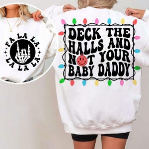 Puede incluir: Sudadera blanca con un gráfico negro. La parte delantera tiene "FA LA LA" alrededor de una mano de esqueleto. La parte trasera muestra "DECK THE HALLS AND NOT YOUR BABY DADDY" con un borde de luces navideñas coloridas. Un diseño festivo y humorístico.