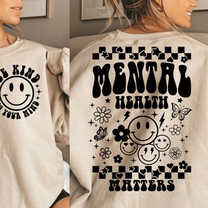 May include: A beige sweatshirt with a black smiley face and the text "Be Kind To Your Mind" on the front and the text "Mental Health Matters" with a black smiley face and other black graphic elements on the back.