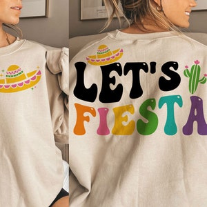 May include: A beige sweatshirt with a colorful "Let's Fiesta" graphic and a small sombrero design. The graphic features a cactus and colorful letters.