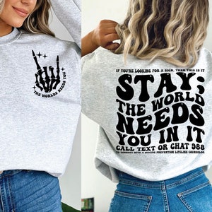 Puede incluir: Sudadera gris claro con texto gr&aacute;fico negro y una mano de esqueleto en la parte delantera. La parte trasera de la sudadera tiene las palabras "STAY; THE WORLD NEEDS YOU IN IT" y un n&uacute;mero de tel&eacute;fono de prevenci&oacute;n del suicidio.