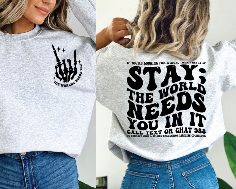 Puede incluir: Sudadera gris claro con texto gr&aacute;fico negro y una mano de esqueleto en la parte delantera. La parte trasera de la sudadera tiene las palabras "STAY; THE WORLD NEEDS YOU IN IT" y un n&uacute;mero de tel&eacute;fono de prevenci&oacute;n del suicidio.