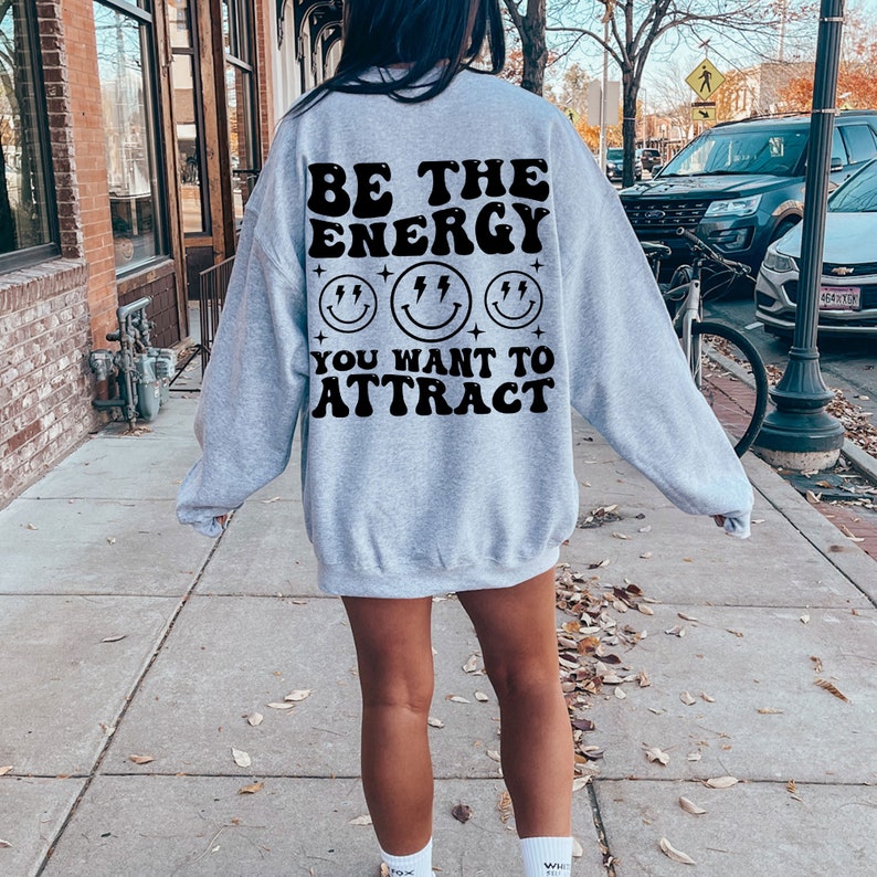 K&ouml;nnte beinhalten: Grauer Pullover mit einem schwarzen Aufdruck, der "Sei die Energie, die du anziehen m&ouml;chtest" mit drei Smiley-Gesichtern und Sternen.