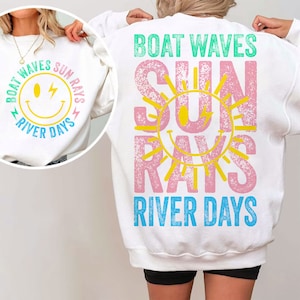 Op de afbeelding: Wit sweatshirt met een roze en gele zon graphic en de tekst "BOAT WAVES SUN RAYS RIVER DAYS".