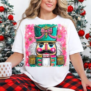 K&ouml;nnte beinhalten: Wei&szlig;es T-Shirt mit einer farbenfrohen Illustration eines Nussknackersoldaten. Das Design zeigt einen t&uuml;rkisfarbenen Hut mit einer rosa Schleife, einen rosa Hintergrund mit goldenen Sternen und einen schwarzen Schnurrbart. Weihnachtsthema.