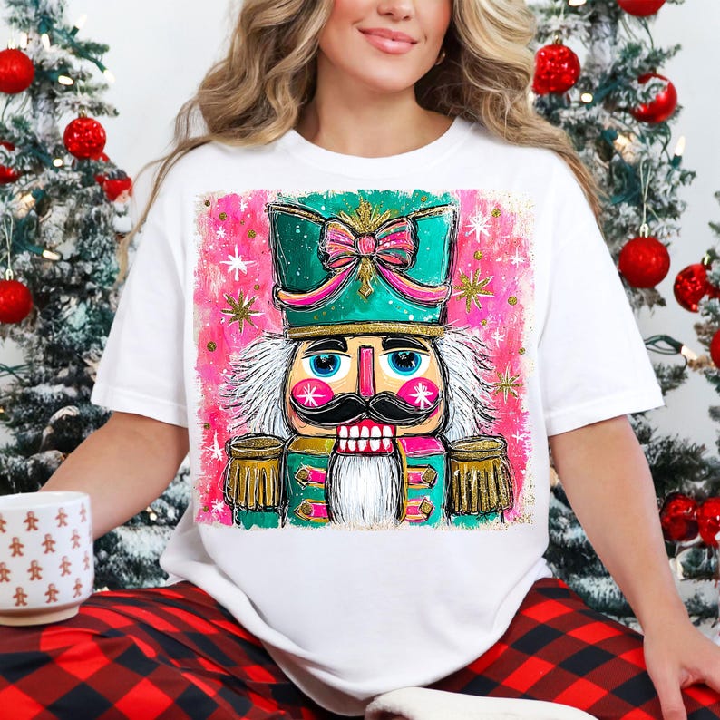 K&ouml;nnte beinhalten: Wei&szlig;es T-Shirt mit einer farbenfrohen Illustration eines Nussknackersoldaten. Das Design zeigt einen t&uuml;rkisfarbenen Hut mit einer rosa Schleife, einen rosa Hintergrund mit goldenen Sternen und einen schwarzen Schnurrbart. Weihnachtsthema.
