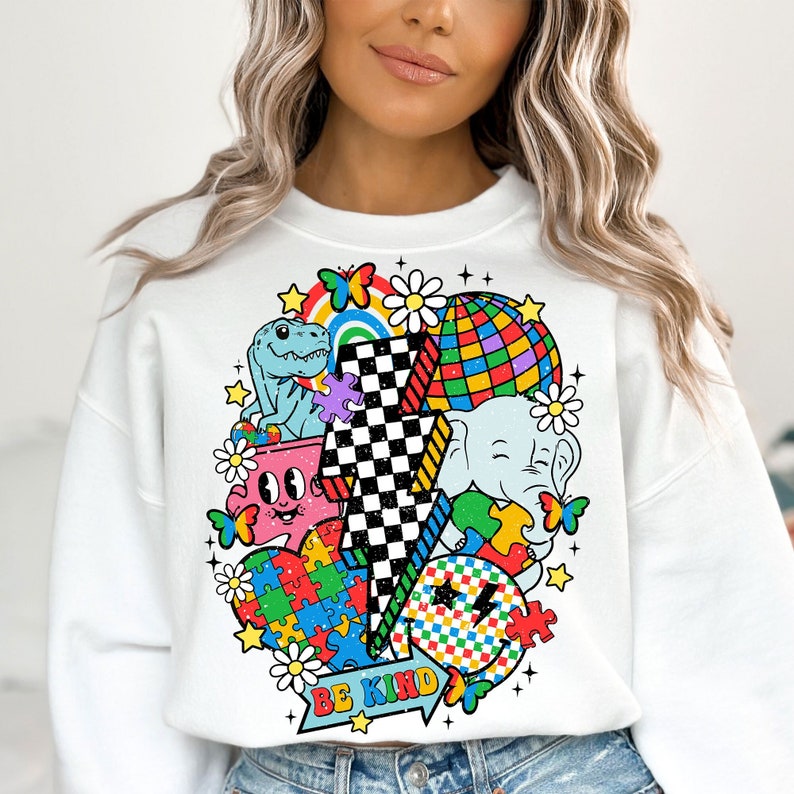 Op de afbeelding: Witte trui met een kleurrijke grafische print met een bliksem, een dinosaurus, een olifant, een regenboog, een puzzelstuk, een madeliefje en een discobal. De grafiek bevat ook de tekst "BE KIND".
