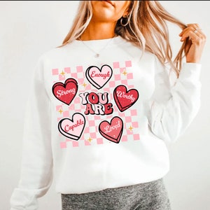 May include: White sweatshirt with a pink and white checkered background and red hearts with positive affirmations. The hearts say "Strong", "Enough", "Worthy", "Capable", and "Loved". The text "You Are" is in large, pink letters.