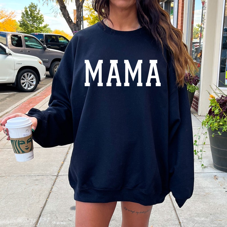 May include: A black sweatshirt with the word "MAMA" printed in white on the front. The sweatshirt is being worn by a person holding a coffee cup.