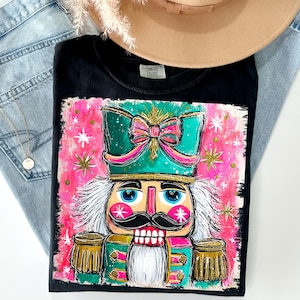 K&ouml;nnte beinhalten: Schwarzes T-Shirt mit einer farbenfrohen Darstellung eines Nussknackers. Das Design umfasst eine t&uuml;rkisgr&uuml;ne M&uuml;tze mit rosa Schleife, einen wei&szlig;en Bart und goldene Akzente. Das T-Shirt liegt auf einer Jeans und einem beigen Hut.