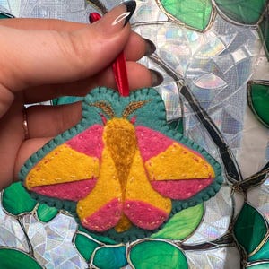 May include: A handmade felt moth ornament with a teal body, pink and yellow wings, and gold antennae. The ornament has a red ribbon for hanging. The moth is a decorative item.