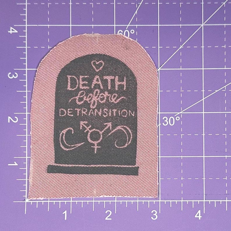 May include: A pink fabric patch in the shape of a tombstone. The patch has the words "DEATH Before DETRANSITION" in white script, along with a heart and gender symbols. The patch is approximately 2.5 inches tall and 2 inches wide.