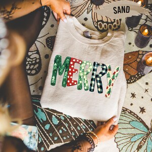 May include: A sand-colored sweatshirt with the word "MERRY" in colorful, textured letters. The letters are green, red with white polka dots, white with green polka dots, black and white plaid, and a festive pattern with Christmas trees. The sweatshirt is folded.