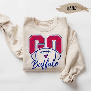 May include: A sand-colored sweatshirt with the words "GO Buffalo" in a red and blue design. The word "GO" is in large block letters with a patterned fill, and the word "Buffalo" is written in script below a football graphic.