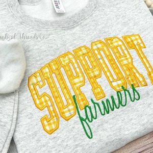 May include: A light gray sweatshirt with the words "SUPPORT Farmers" embroidered in yellow plaid and green thread. The sweatshirt has a crew neck and long sleeves. The brand name "Jugbird Threads Co." is printed on the left side.