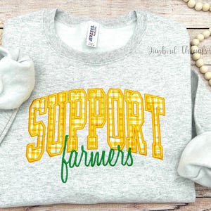 May include: A light gray sweatshirt with the words "SUPPORT farmers" in yellow plaid lettering and green script. The sweatshirt has long sleeves and a crew neck. The image is on a light wood background.