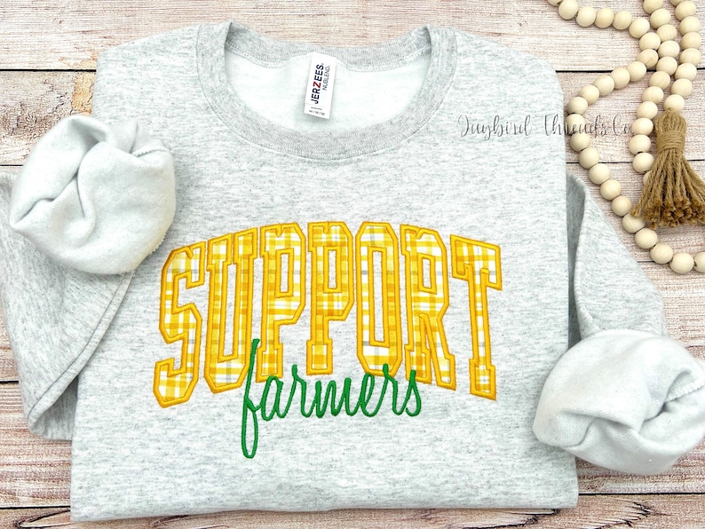 May include: A light gray sweatshirt with the words "SUPPORT farmers" in yellow plaid lettering and green script. The sweatshirt has long sleeves and a crew neck. The image is on a light wood background.