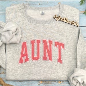 May include: A light gray sweatshirt with the word "AUNT" in a red, glittery, collegiate-style font. The sweatshirt has a crew neck and long sleeves. The brand name "Joybird Threads Co." is on a wooden tag.