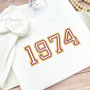 May include: White sweatshirt with a gold and maroon embroidered design of the year 1974.