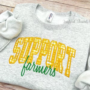 May include: A light gray sweatshirt with the words "SUPPORT farmers" in yellow plaid lettering and green script. The sweatshirt has a crew neck and long sleeves. The brand tag is visible.