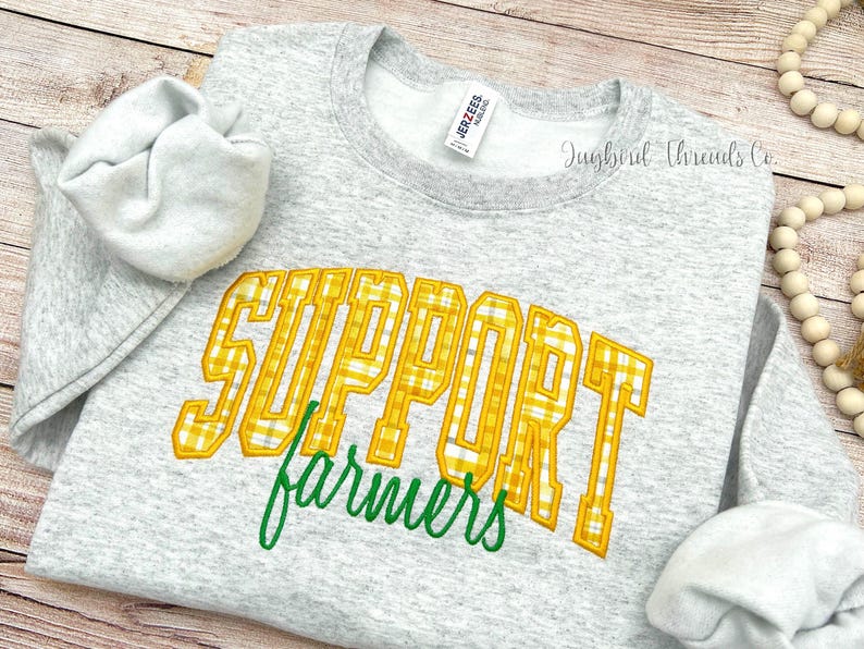 May include: A light gray sweatshirt with the words "SUPPORT farmers" in yellow plaid lettering and green script. The sweatshirt has a crew neck and long sleeves. The brand tag is visible.