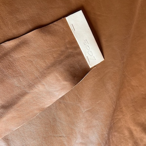 Könnte beinhalten: Nahaufnahme eines Stücks braunen Leders mit glatter Textur. Ein kleines, rechteckiges Etikett mit der Nummer "5007" ist auf dem Leder befestigt. Das Leder hat eine warme, natürliche Farbe und scheint hochwertig zu sein.