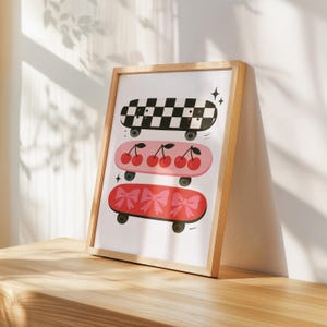 May include: A framed print featuring three skateboards with unique designs. The top skateboard has a black and white checkered pattern. The middle skateboard is pink with cherries. The bottom skateboard is red with pink bows. The frame is made of light-colored wood.