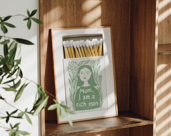 May include: A white wooden box with a green illustration of a woman with the text "Mom, I am a rich man" inside. The box contains a stack of yellow matches with white tips.