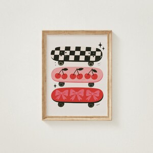 May include: A framed print featuring three skateboards stacked vertically. The top skateboard has a black and white checkerboard pattern. The middle skateboard is pink with cherries. The bottom skateboard is red with bows.