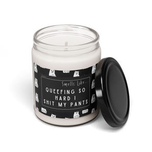 May include: A white candle in a glass jar with a black lid. The candle has a label with a black background and white text that reads "Smells Like... QUEEFING SO HARD I SHIT MY PANTS". The label also features a pattern of white cat illustrations.