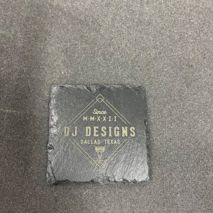 May include: A black slate coaster with gold lettering that reads "Since M-M-X-X-I-I DJ DESIGNS DALLAS TEXAS".