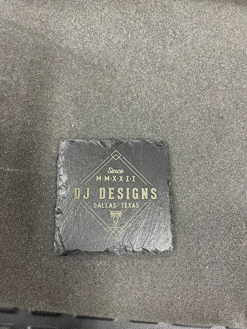 May include: A black slate coaster with gold lettering that reads "Since M-M-X-X-I-I DJ DESIGNS DALLAS TEXAS".