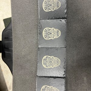 May include: Set of four black slate coasters with a white sugar skull design. Each coaster features a different skull design with intricate details.