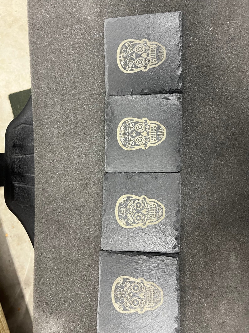 May include: Set of four black slate coasters with a white sugar skull design. Each coaster features a different skull design with intricate details.