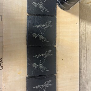 May include: Four black slate coasters featuring a laser-etched design of two hummingbirds facing each other. The hummingbirds are in a light grey colour.