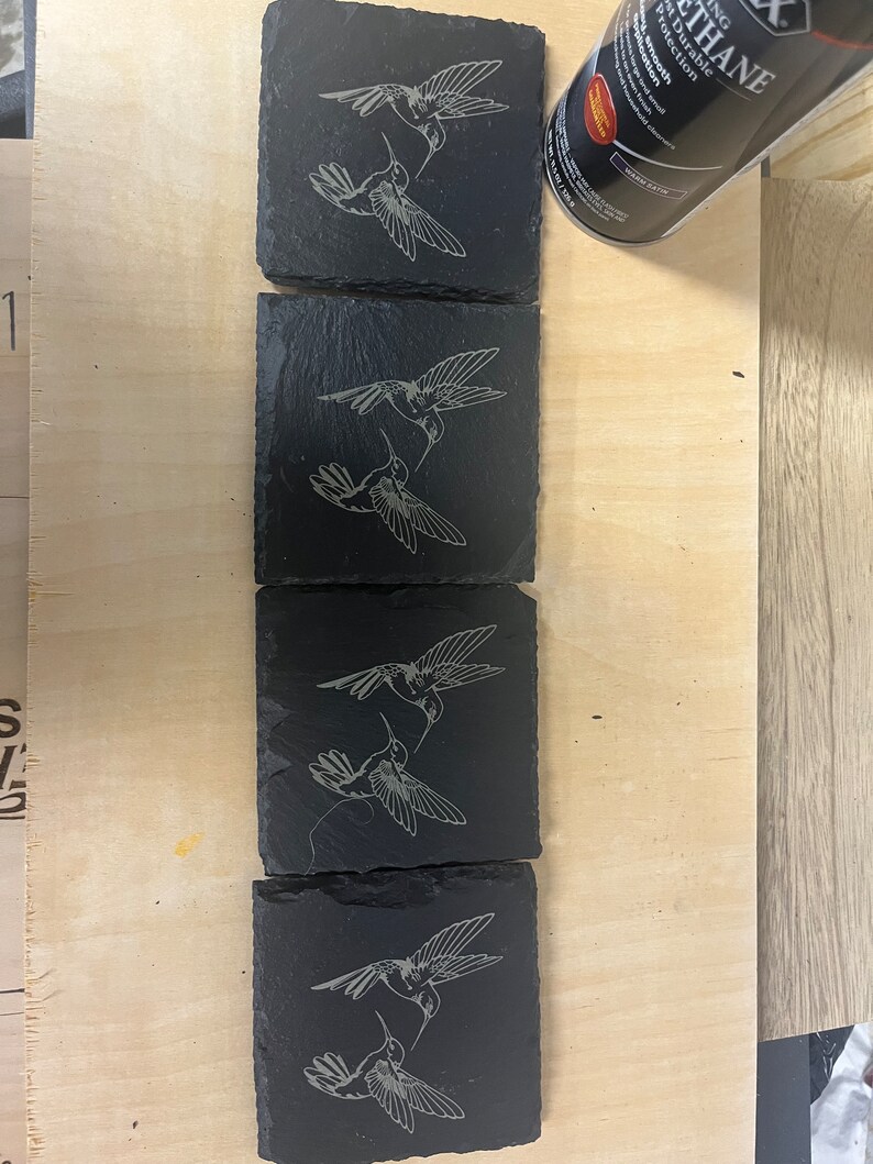 May include: Four black slate coasters featuring a laser-etched design of two hummingbirds facing each other. The hummingbirds are in a light grey colour.