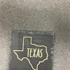 May include: A black slate coaster with a gold outline of the state of Texas and the word "TEXAS" written inside the outline.