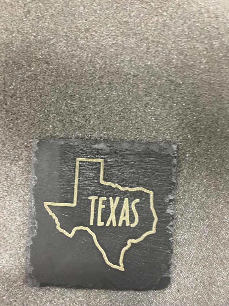May include: A black slate coaster with a gold outline of the state of Texas and the word "TEXAS" written inside the outline.