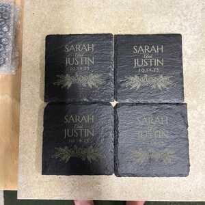 May include: Four black slate coasters with gold lettering. The coasters are engraved with the names "Sarah and Justin" and the date "10.14.23".